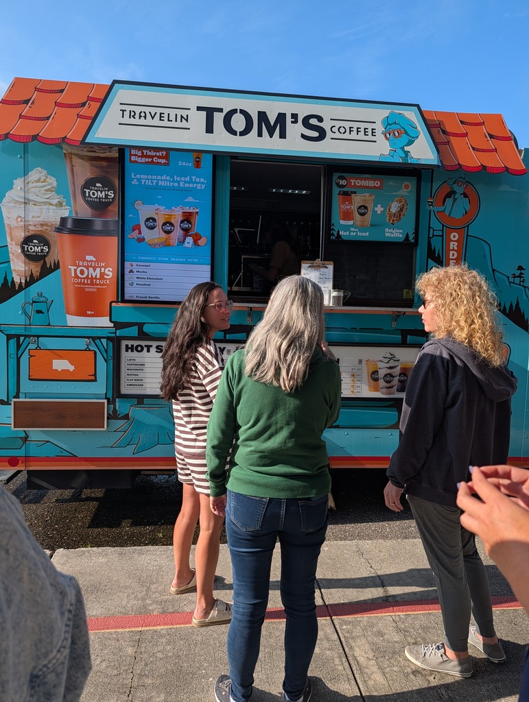Three women stand in front of a food truck with "Tom's Coffee" on the side. One woman faces the truck, another faces the camera.