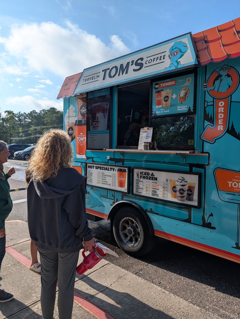 A person stands in front of a coffee truck. The truck has blue and orange colors and is parked on the street.