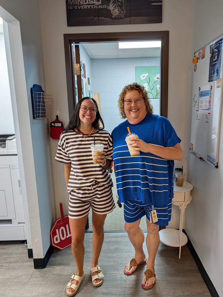 Two people in a room, one wearing a striped shirt and shorts, the other in a blue shirt and shorts. They hold drinks.