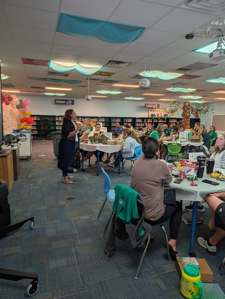 A group of people are seated around tables in a room with bookshelves, balloons, and a projector screen.