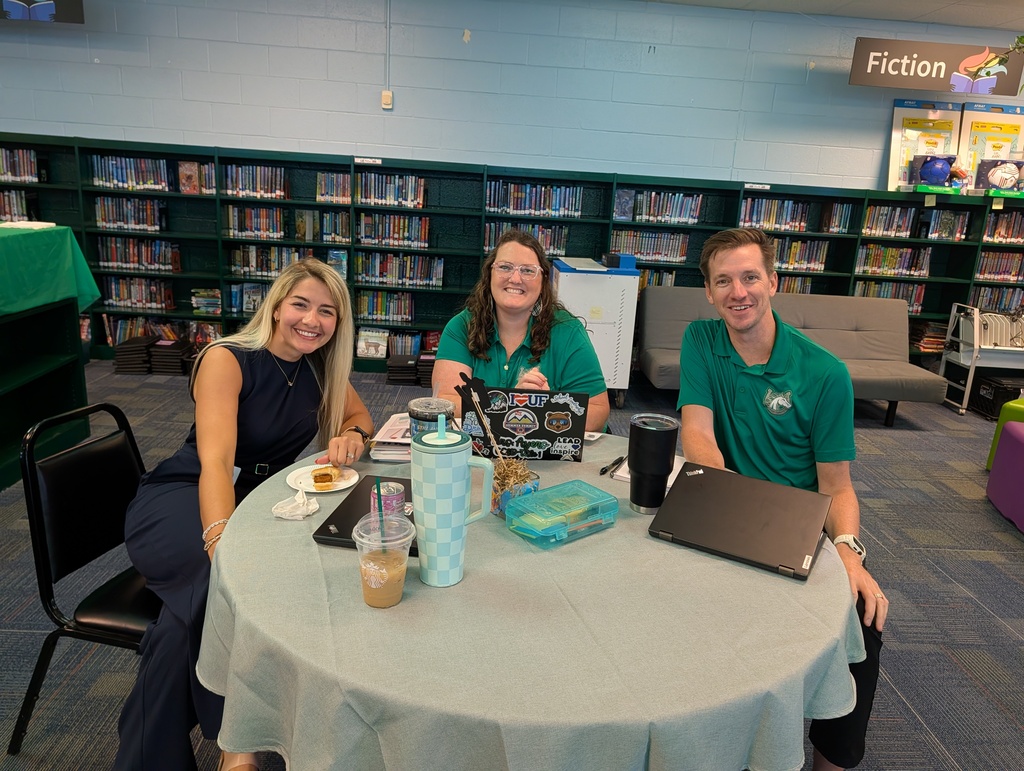 Three people in green shirts sit at a round table in a library. A woman and two men smile.