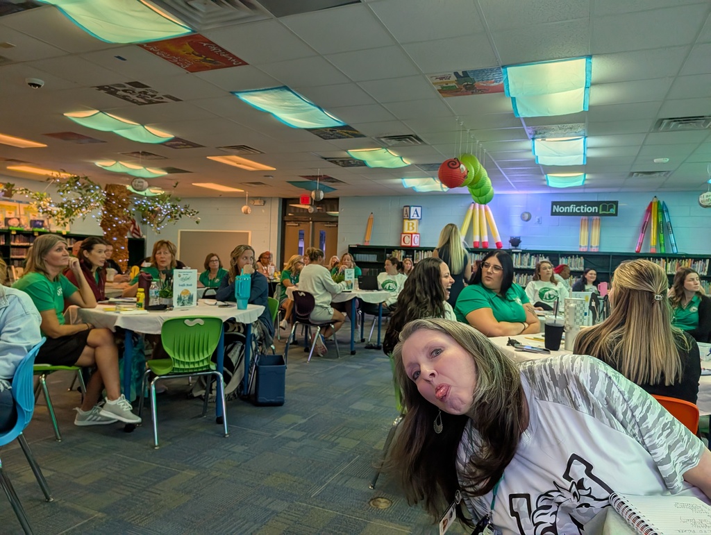 Several people in green shirts sit at tables in a room with lamps and balloons. A woman with long dark hair rests her head on a table.