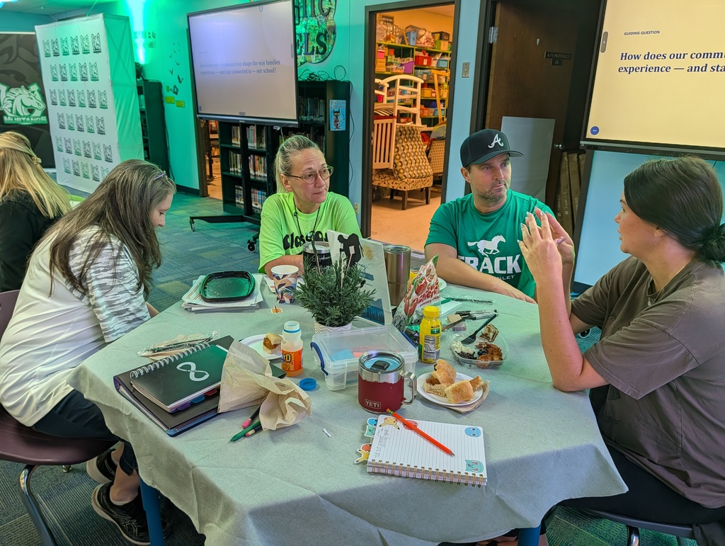 Four people sit at a table with books, a plant, and food. A projector screen is in the background.