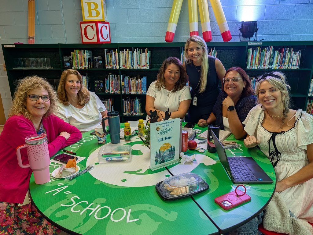 Group of women in dresses sitting around a green table with the word "School" on it.
