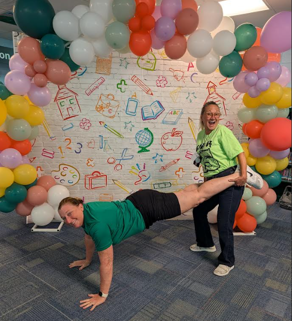 A person performs a plank while another holds their leg in a room with colorful balloons and a mural.