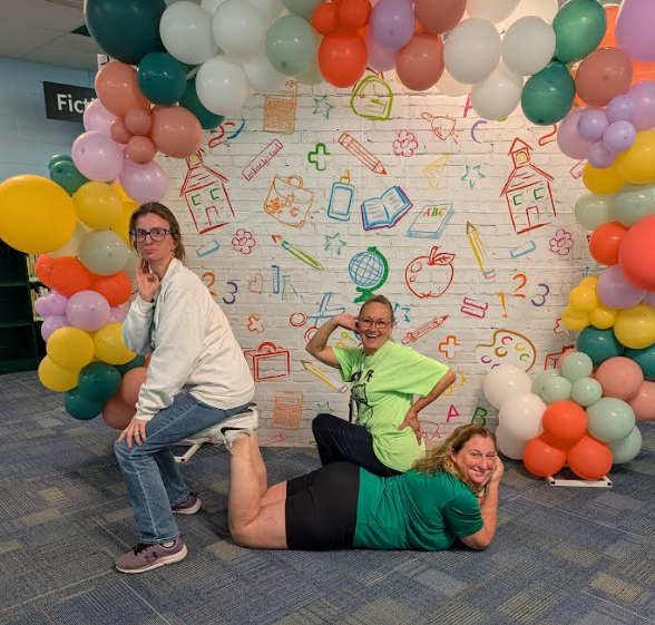 Three people pose for a photo, two sitting and one kneeling, in front of a colorful balloon arch and a mural with school-themed drawings.
