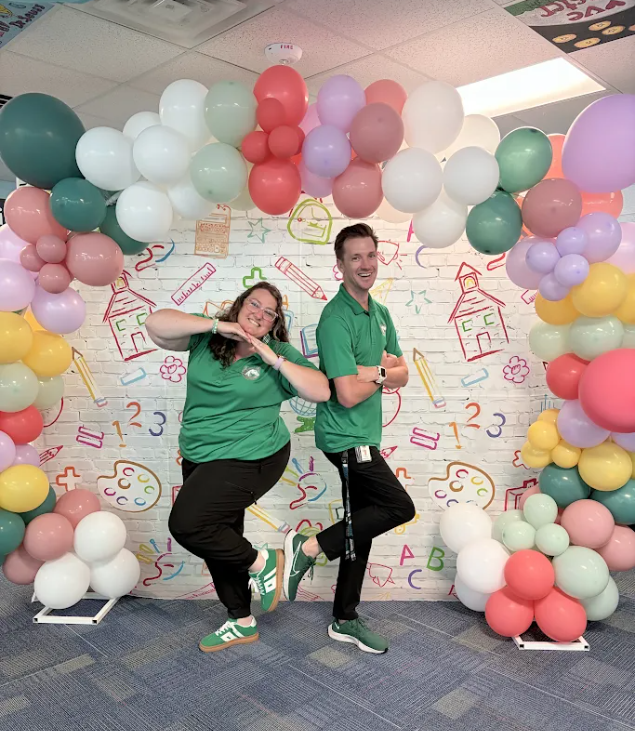 Two people wearing green shirts and black pants pose in front of a colorful balloon arch.