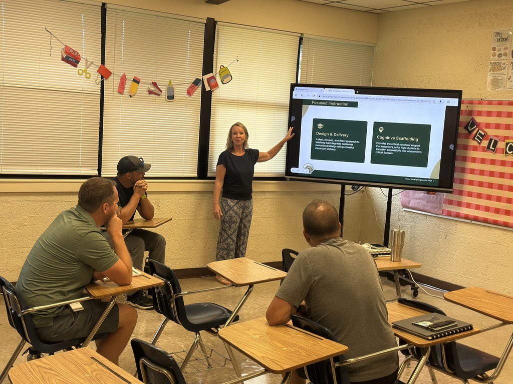 A classroom with a large screen. A woman points at the screen. Four students sit at desks.