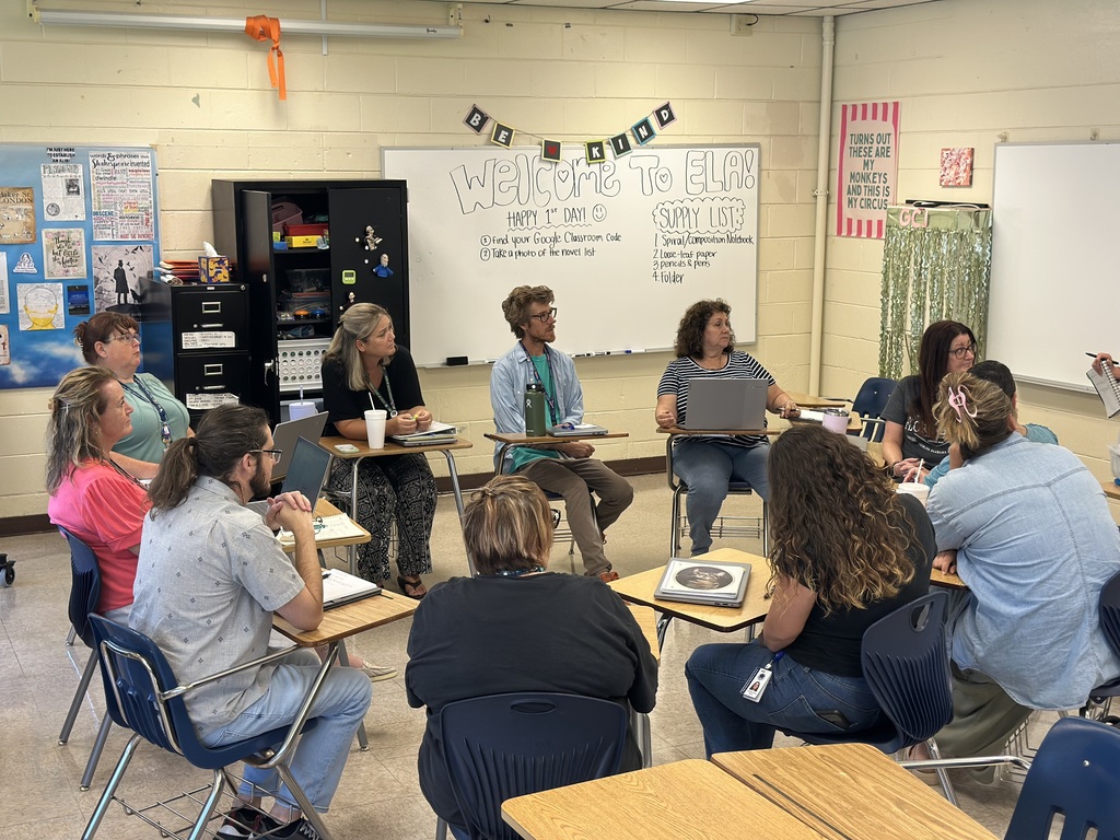 A classroom with desks and chairs, a whiteboard with writing, and people sitting in chairs, facing each other.