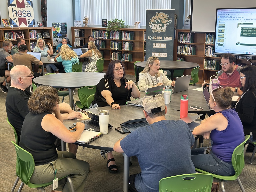 Library meeting with people at tables. A screen is displayed on a wall. Bookshelves are behind them.