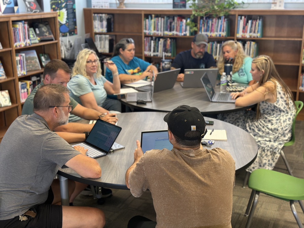 A group of people sit around tables with laptops in a library, discussing with each other.