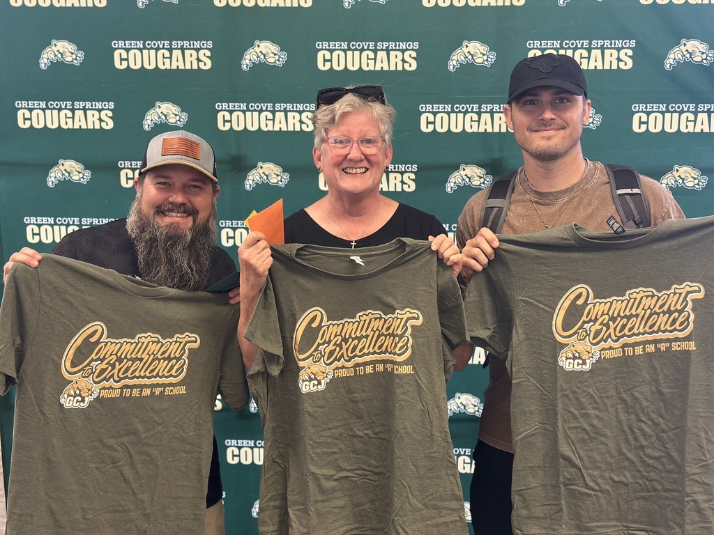 Three people holding green shirts with the words "Commitment Excellence" in front of a green wall with team logos.