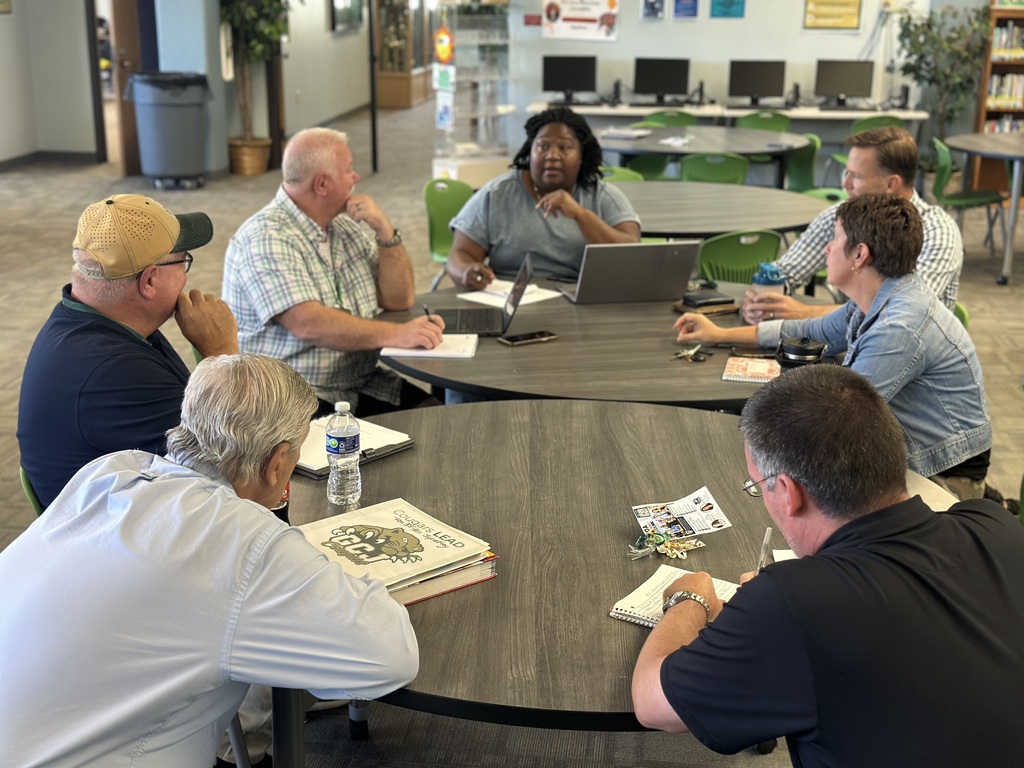 A group of people sit around a table in a room with computers and plants.