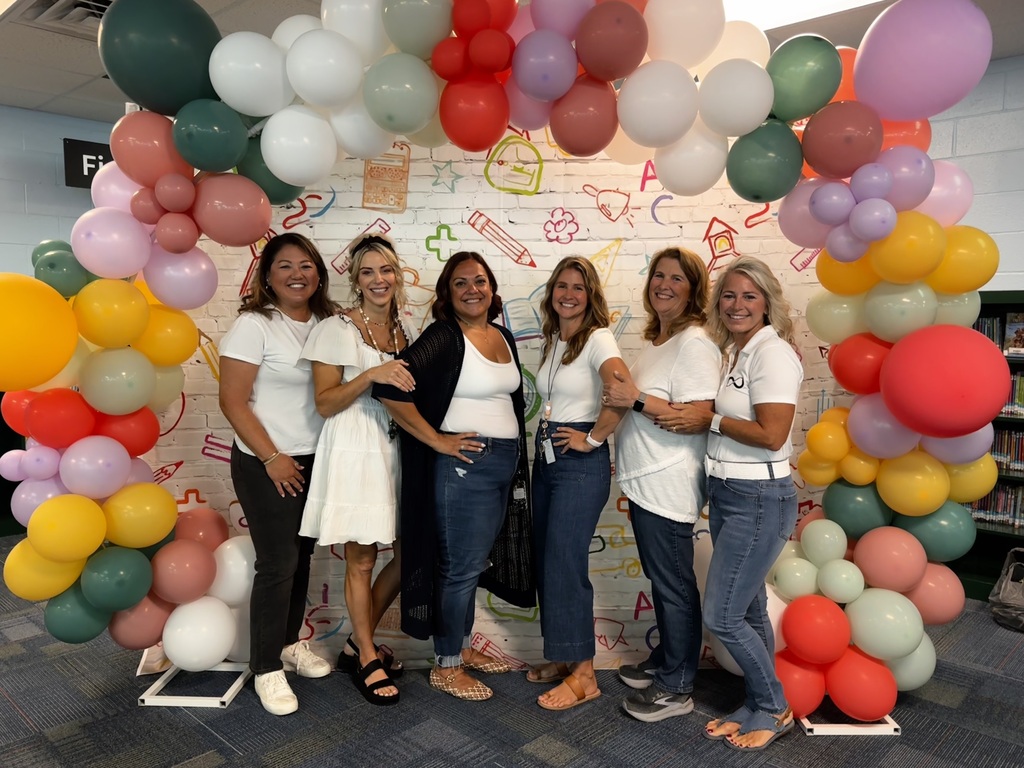 Five women in white shirts stand under a colorful balloon arch in a room.