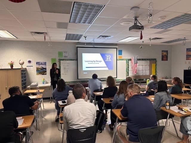 People sit in chairs at desks in a classroom. A woman stands before a projector screen displaying text.
