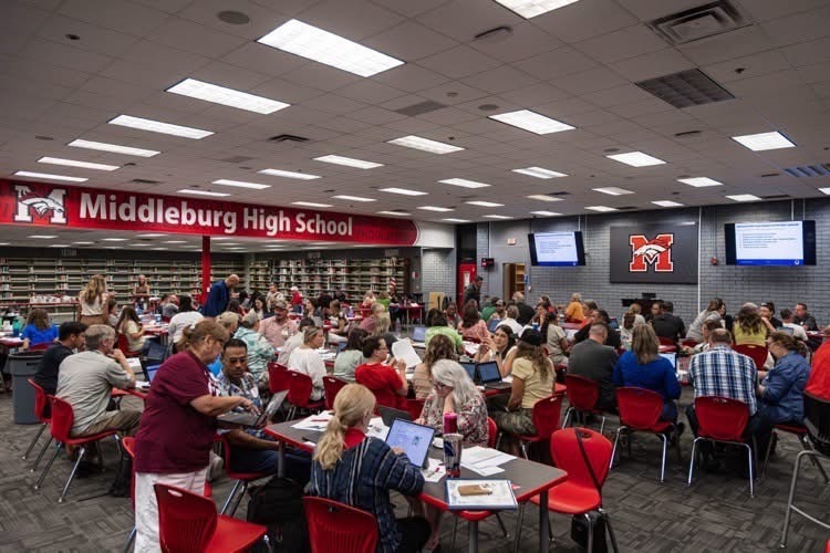 A large group of people seated at tables in a room. Bright overhead lights. A sign on the wall says "Middleburg High School."