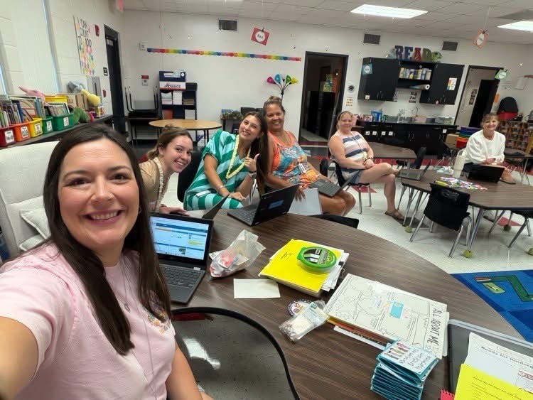 A group of smiling adults in a classroom with a table, laptops, and papers.