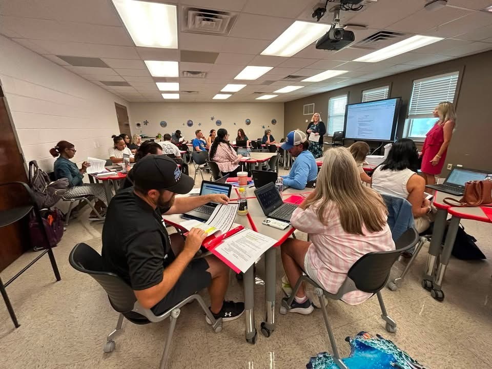 A classroom with desks. Adults sit around tables, some with laptops. A woman stands at a screen.