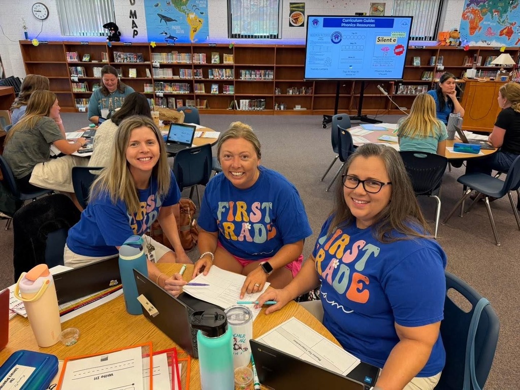 Three women wear blue shirts with the word "First" in front of a library table.
