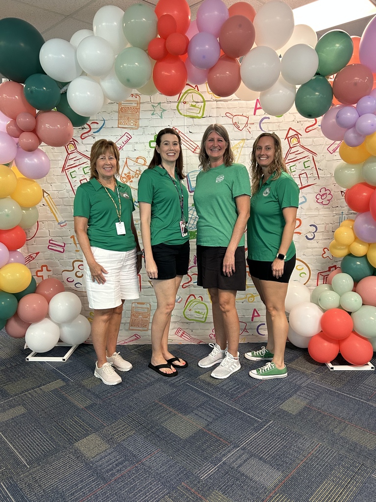 Four women in green t-shirts stand in a colorful balloon arch. The wall behind them has hand-drawn illustrations.