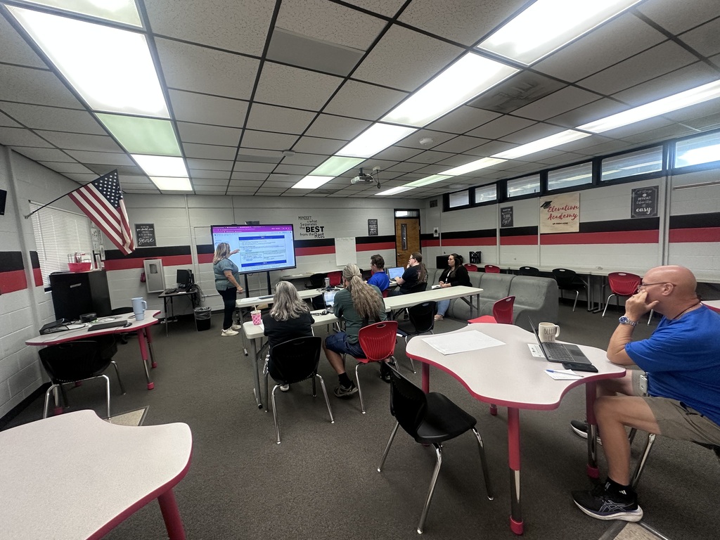 Classroom with white tables, chairs, and a person seated at a desk with a laptop.