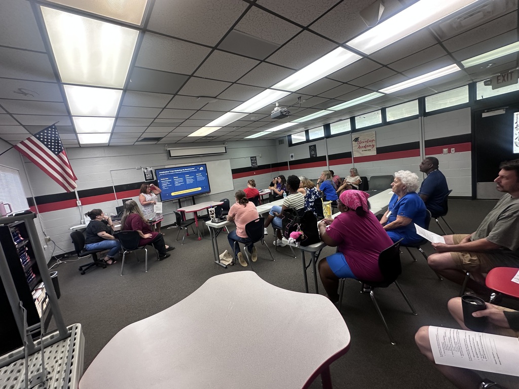 A group of people sit in chairs around tables in a classroom. A projector screen is on the wall.
