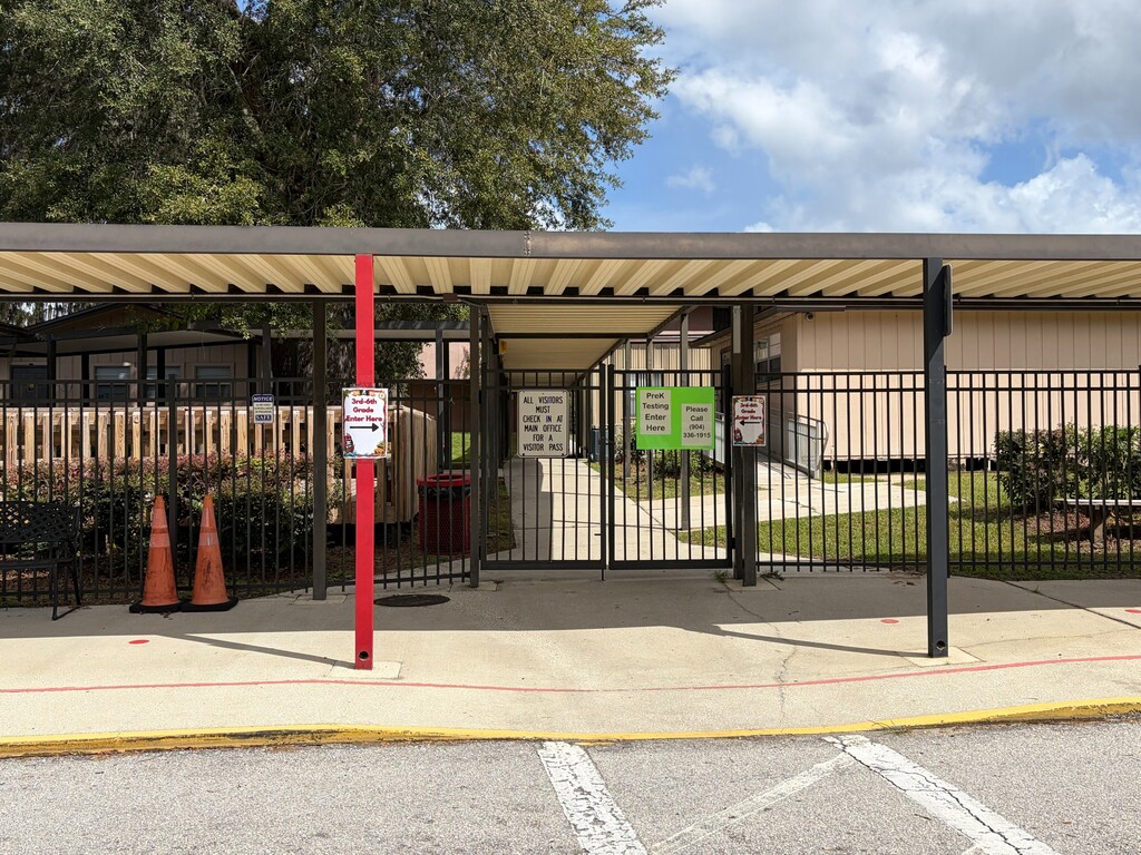 Entrance to a building with a metal roof, fence, and gate. Two orange traffic cones and a green sign are nearby.