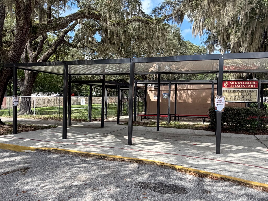 A shaded outdoor waiting area with a metal frame roof, benches, and a red sign. Trees surround the area.