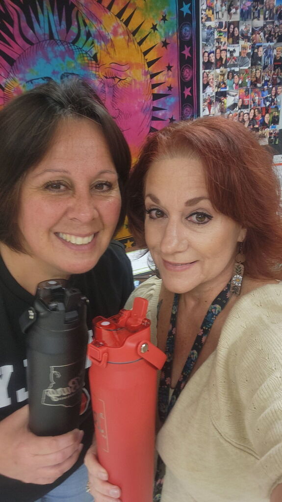 Two women smiling, one holding a black tumbler and the other holding a red tumbler, in front of a colorful wall.