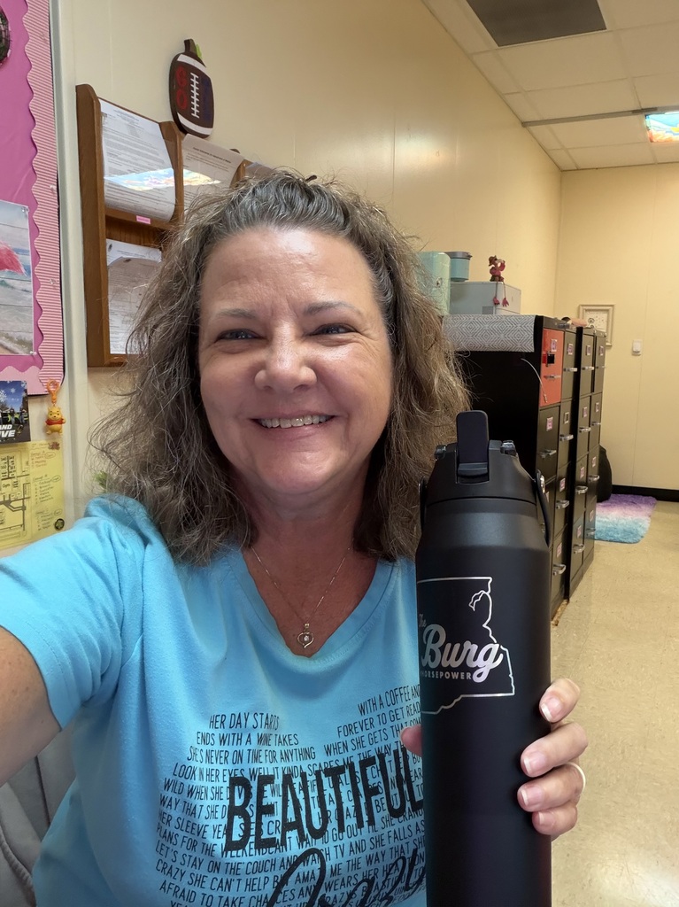 A woman in a blue shirt takes a selfie holding a black tumbler. The background includes a classroom with cabinets and posters.