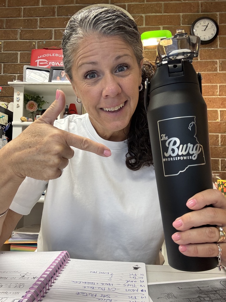 A woman in a white t-shirt is holding a black tumbler and pointing. Behind her is a brick wall, shelves, a clock, and a bright green lamp.