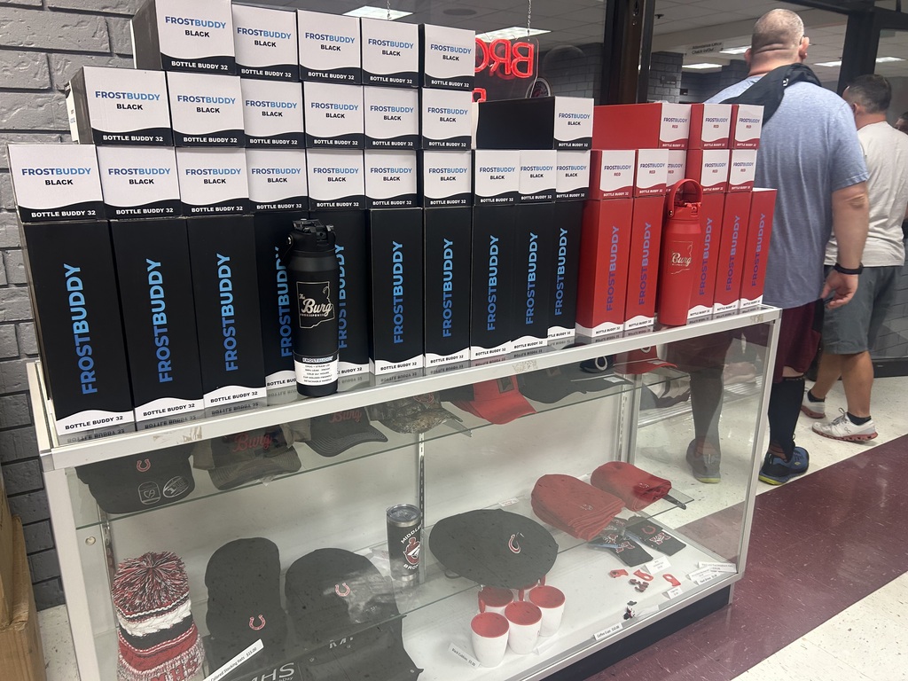 A display case holds various items including black and red boxes, a water bottle, hats, cups, and a storage box. A man stands nearby.