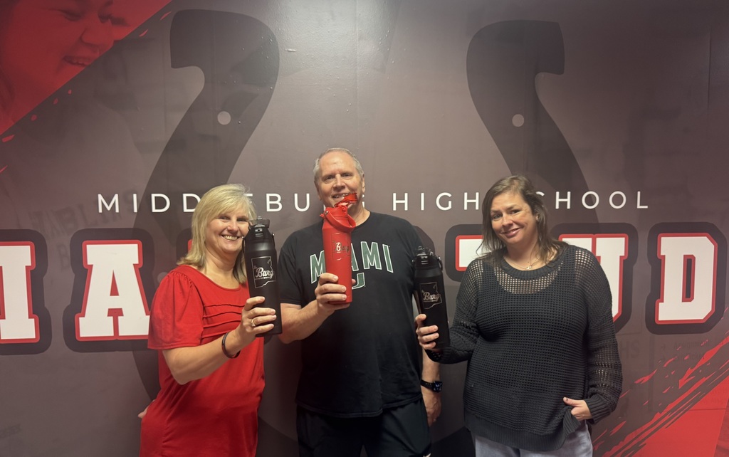 Three people pose for a photo holding water bottles in front of a "MIDDLEBURG  HIGH SCHOOL" sign.