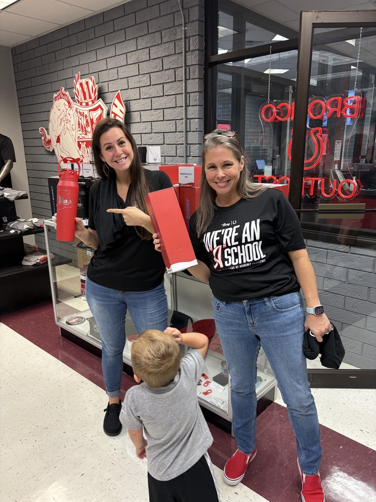 Two women with a child, one holding a cup, stand in a room with shelves, brick walls, and a glass door.