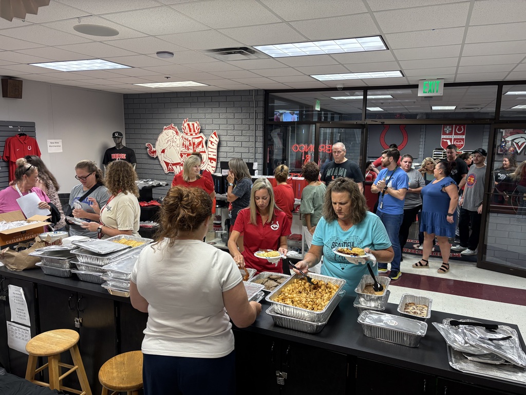 A group of people, including women in white and red shirts, gather around a buffet table indoors.