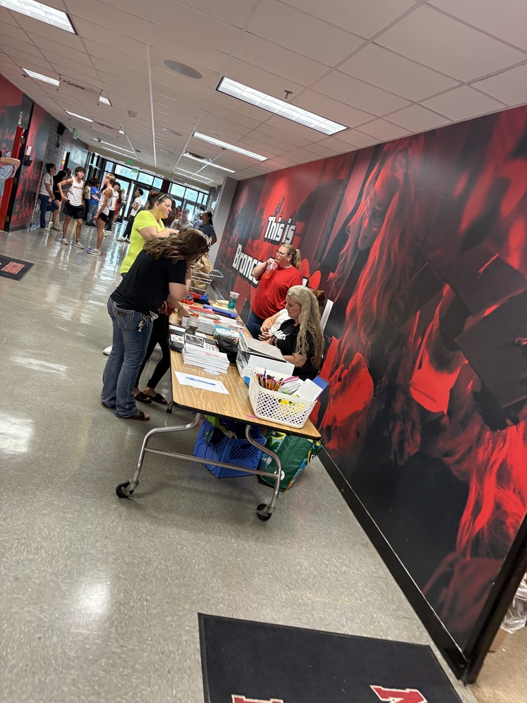 A hallway with a red and black wall mural, people standing, tables, and a rug on the floor.