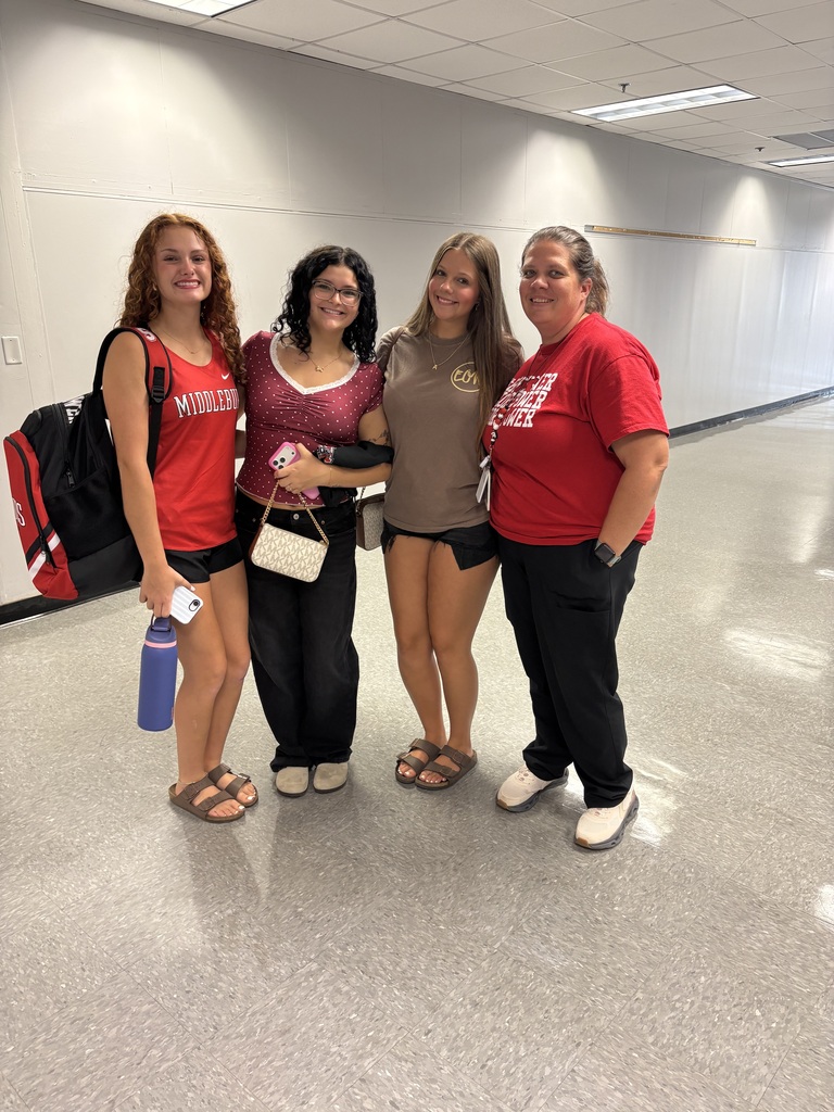Four women in red and brown tops pose for a photo in a school hallway. One holds a water bottle, and one wears a backpack.