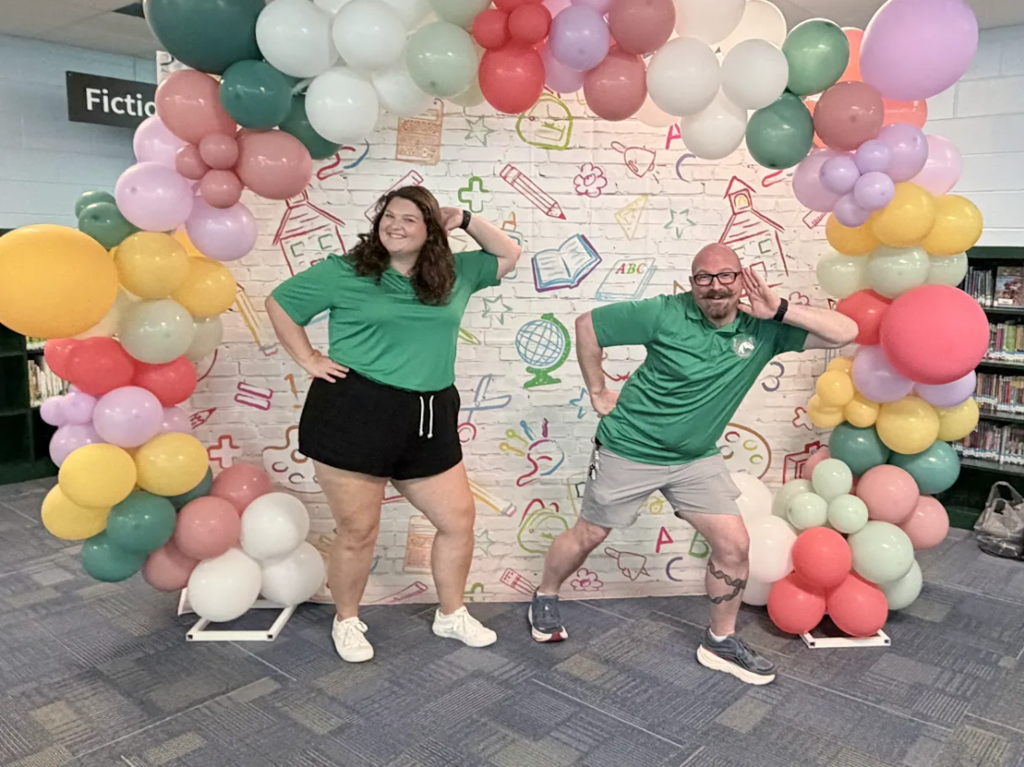 Two individuals wearing green shirts and shorts standing in front of a colorful balloon arch.