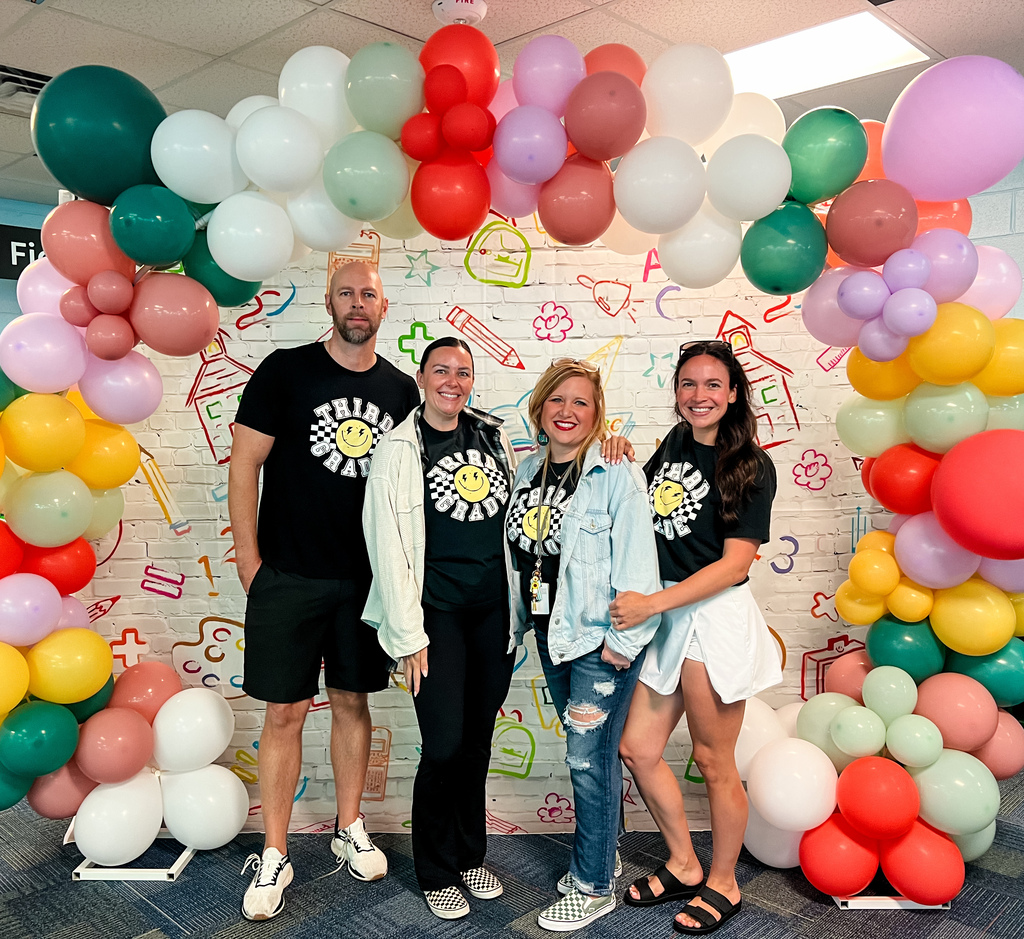 Four people stand behind a colorful balloon arch in a room with a white wall.