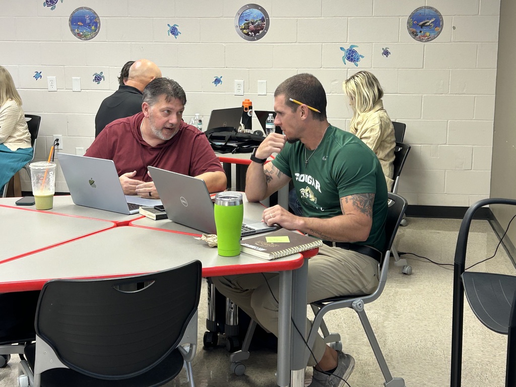 Two men are seated at a table, working on laptops. One wears a red shirt, the other a green shirt. Items on the table include a cup, tumbler, and books.