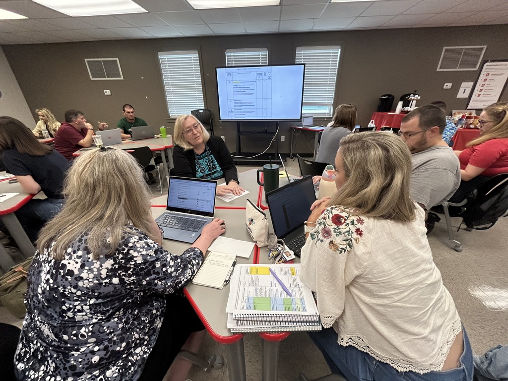 People in a classroom sit at tables with laptops. A woman in a floral shirt speaks to two other women.