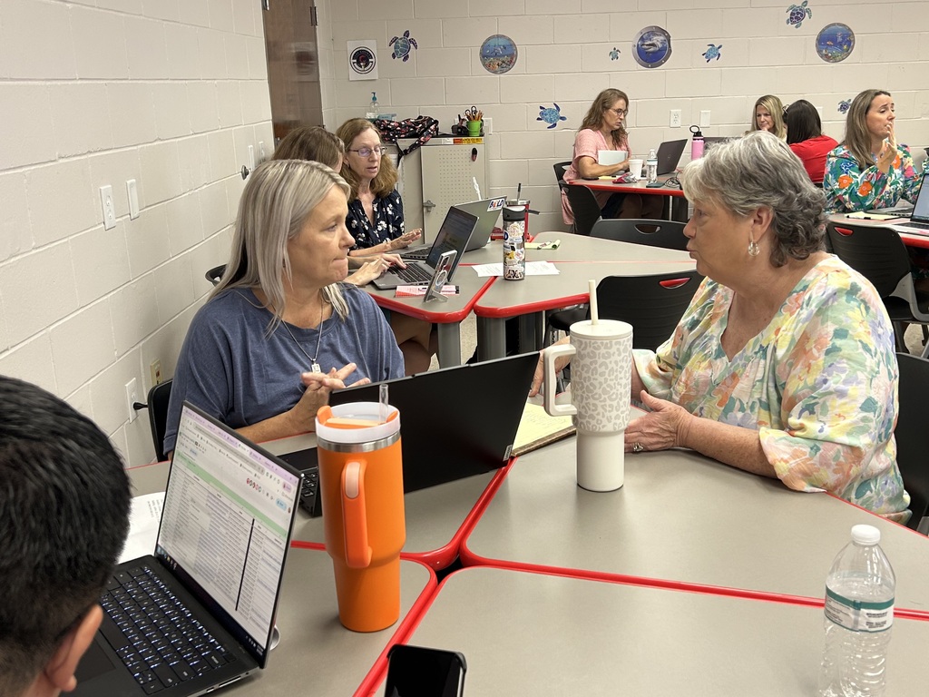 A group of people seated around tables in a room with laptops, one older woman speaks with another.
