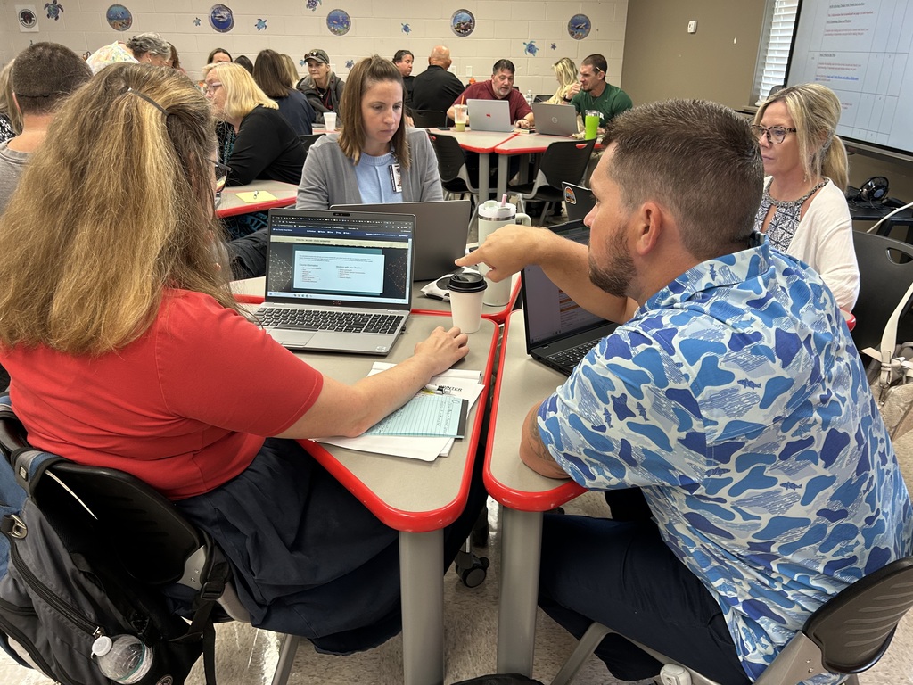 Multiple people sit around a table with laptops, discussing. A projector screen is in the background.