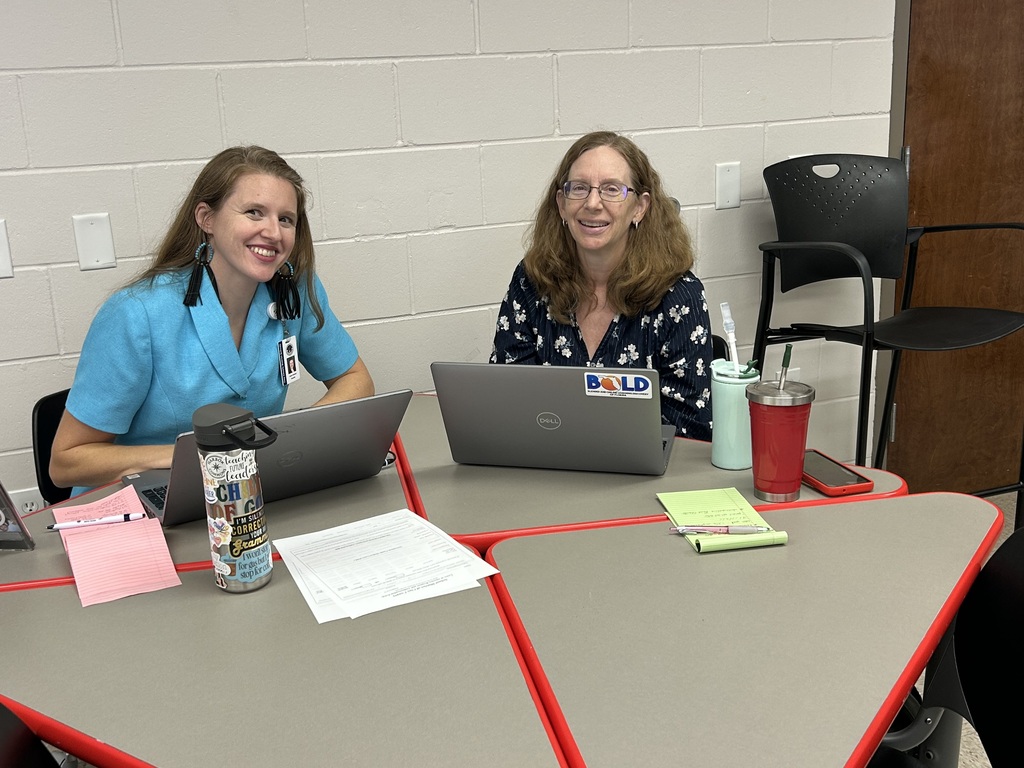 Two women seated at a table with laptops, papers, a tumbler, and a red cup.