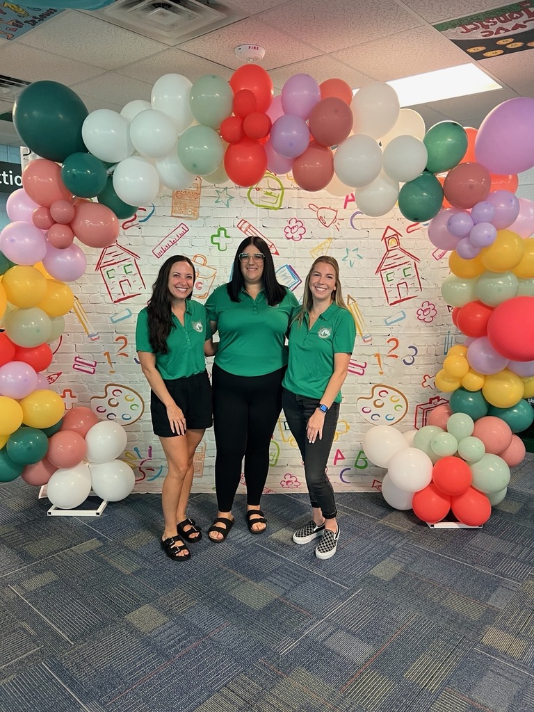 Three women in green shirts and black pants stand under a colorful balloon arch with an educational wall in the background.