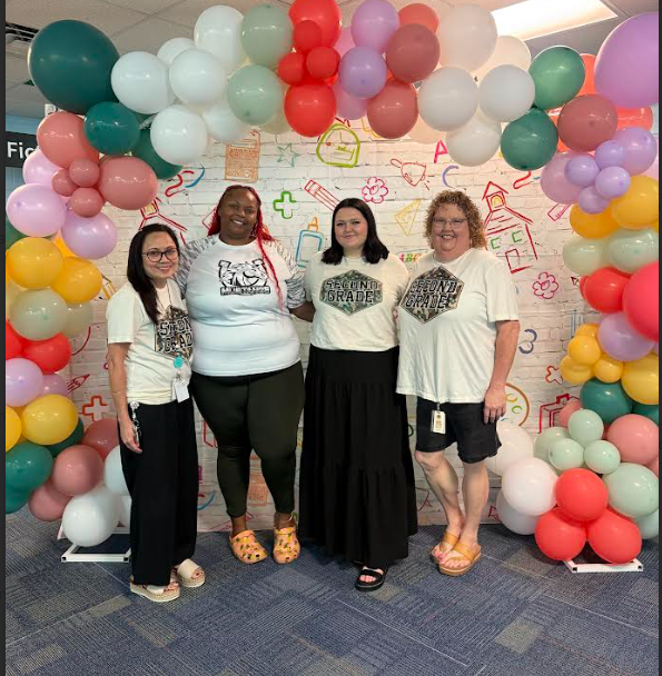 Four women in matching white T-shirts stand under a colorful balloon arch. The background features a white wall with various designs.