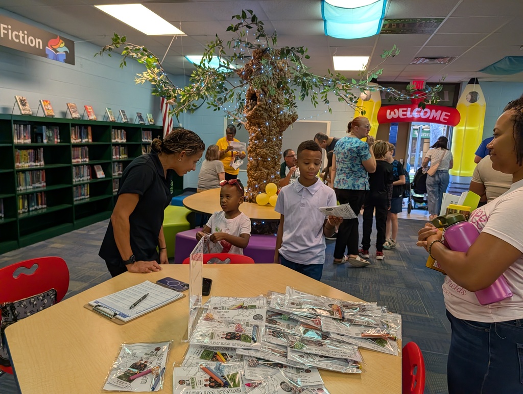 In a library, a woman helps a child at a table with a plant. Nearby, people are walking, and a man reads a book.