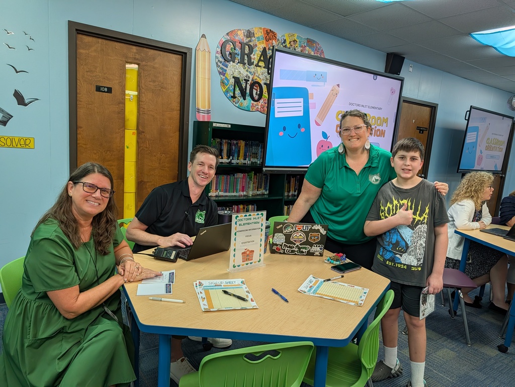 Four people, including a boy, in green and black shirts sit around a table in a classroom. A screen is mounted on the wall.