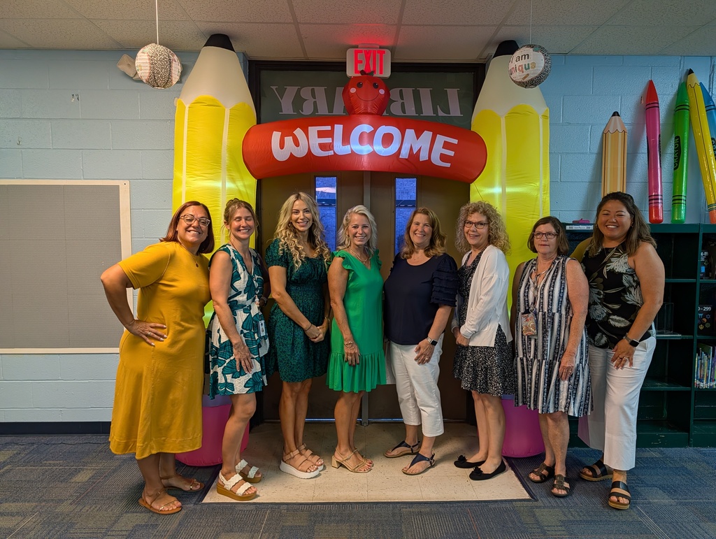 Seven women in various colored dresses stand by a colorful welcome arch in a room. Background includes a white board, shelves, and colorful objects.