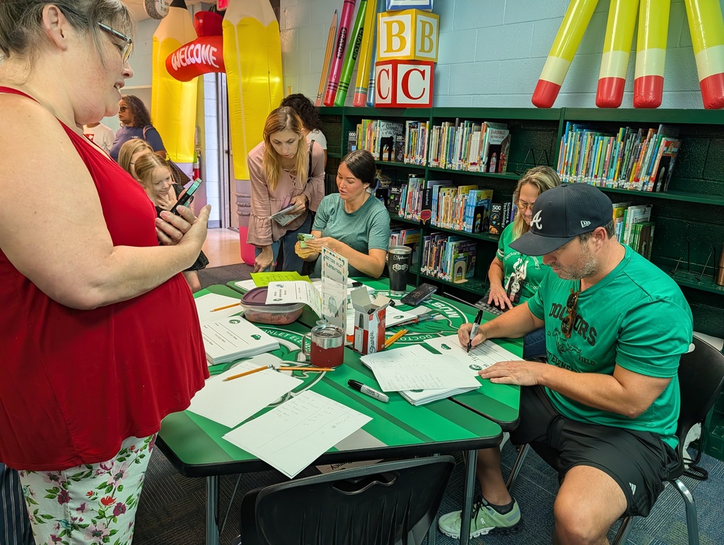 People gather around a table in a library. A woman in red wears glasses. A man in green writes on paper.